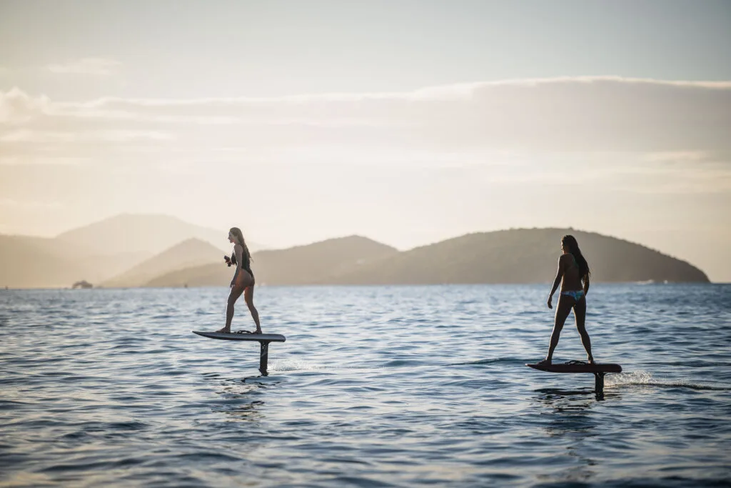 Two people riding eFoil boards at sunset over calm water with distant hills in the background