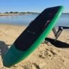 Green electric hydrofoil eFoil board with black deck pad, carbon fiber mast, and foil wings resting on sandy Chesapeake Bay shoreline in Hampton, Virginia.