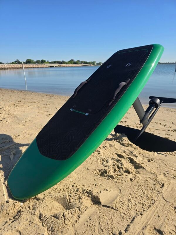 Green electric hydrofoil eFoil board with black deck pad, carbon fiber mast, and foil wings resting on sandy Chesapeake Bay shoreline in Hampton, Virginia.
