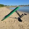 Green electric hydrofoil eFoil board with carbon fiber mast and foil wings resting on the sand beside calm Chesapeake Bay water in Hampton, Virginia.