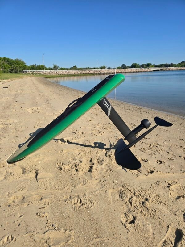 Green electric hydrofoil eFoil board with carbon fiber mast and foil wings resting on the sand beside calm Chesapeake Bay water in Hampton, Virginia.