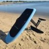 Custom light blue electric hydrofoil eFoil board with black deck pad and foil wings on the sandy shoreline near calm Chesapeake Bay water in Hampton, Virginia.