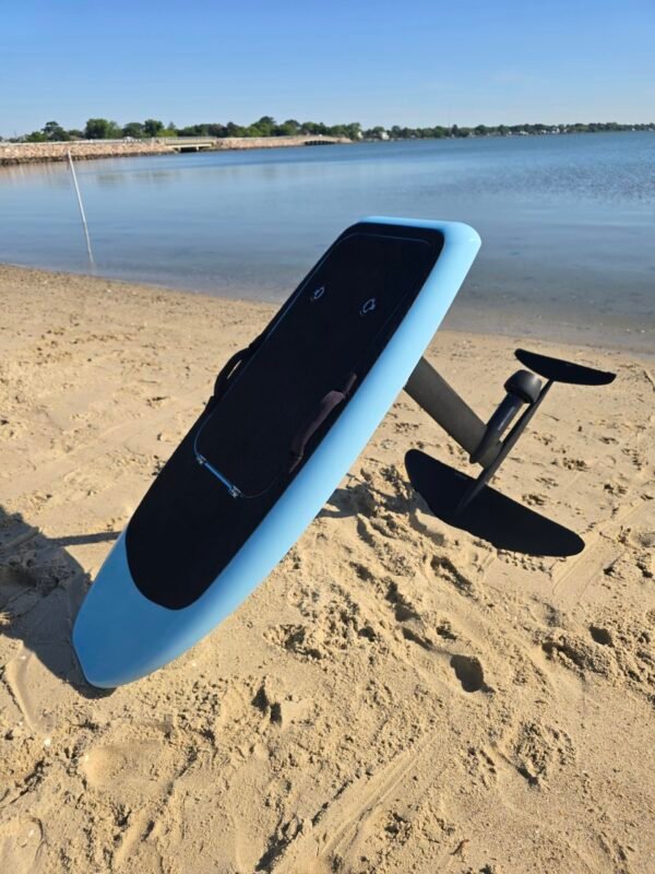 Custom light blue electric hydrofoil eFoil board with black deck pad and foil wings on the sandy shoreline near calm Chesapeake Bay water in Hampton, Virginia.
