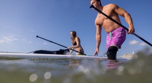 Two people entering the water with paddleboards under a bright blue sky.