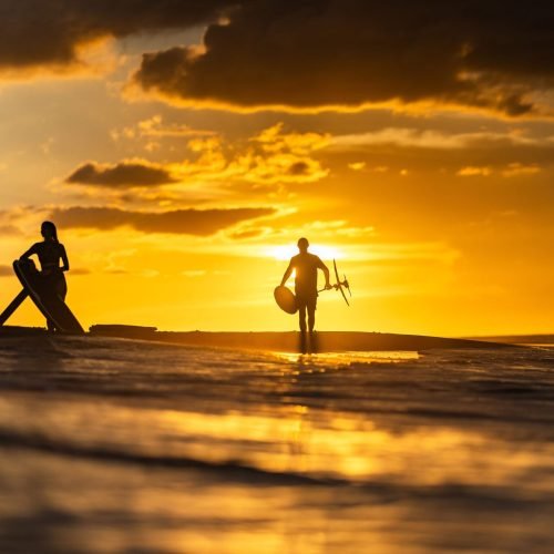 Sunset eFoil Session with LiftX Silhouetted riders with LiftX eFoil boards at the beach during a golden sunset, one carrying an eFoil across the shoreline.
